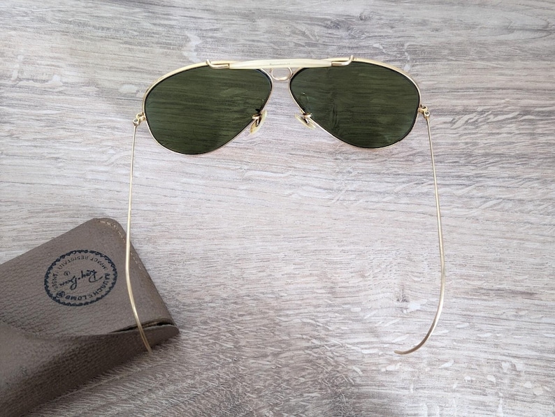 K&ouml;nnte beinhalten: Goldfarbene Sonnenbrille mit gr&uuml;nen Gl&auml;sern. Die Sonnenbrille hat ein klassisches Aviator-Design mit Doppelsteg und d&uuml;nnen B&uuml;geln. Ein braunes Etui mit Logo ist sichtbar.