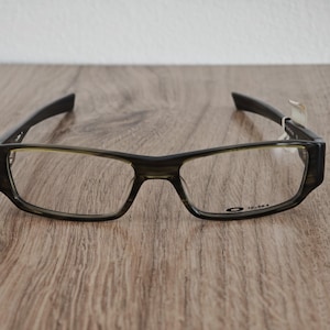 May include: A pair of rectangular-framed eyeglasses with a dark, mottled finish. The frames have a thick, angular design with a subtle green tint around the lenses. The Oakley logo and the number "12-363" are visible on the frame.