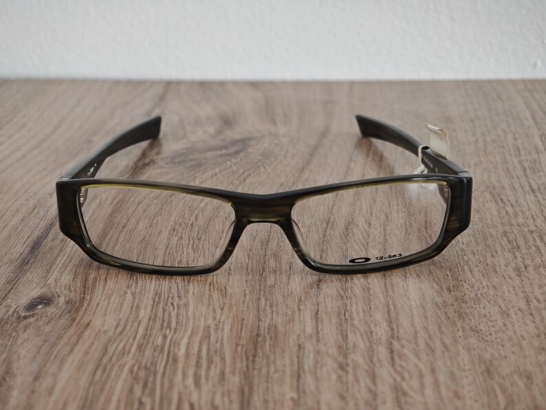 May include: A pair of rectangular-framed eyeglasses with a dark, mottled finish. The frames have a thick, angular design with a subtle green tint around the lenses. The Oakley logo and the number "12-363" are visible on the frame.