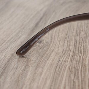 Puede incluir: Una patilla de gafas de color marr&oacute;n transl&uacute;cido con texto impreso en blanco en el lado interior. El texto dice MOD. ET7 G76/198 65 17 125. La patilla de pl&aacute;stico curvada tiene un acabado liso y se estrecha hacia el extremo visible.