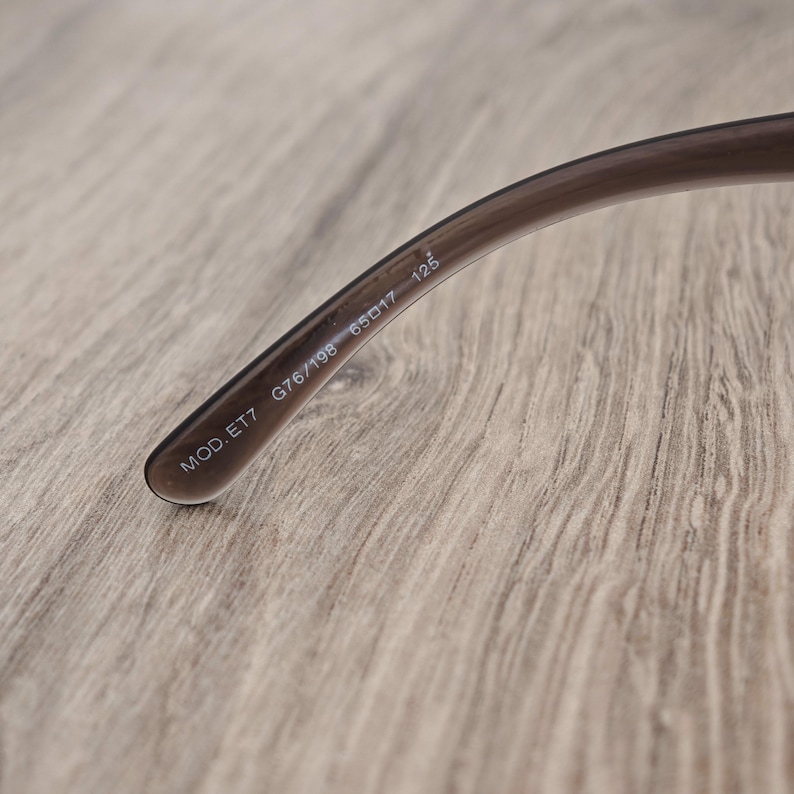 Puede incluir: Una patilla de gafas de color marr&oacute;n transl&uacute;cido con texto impreso en blanco en el lado interior. El texto dice MOD. ET7 G76/198 65 17 125. La patilla de pl&aacute;stico curvada tiene un acabado liso y se estrecha hacia el extremo visible.