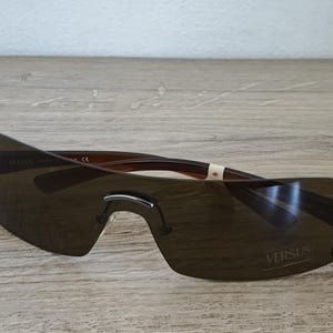 May include: Brown rimless sunglasses with dark tinted lenses. The sunglasses have a curved design and the word "VERSUS" is printed on the lens. The frame is brown and the lenses are a dark shade of brown.