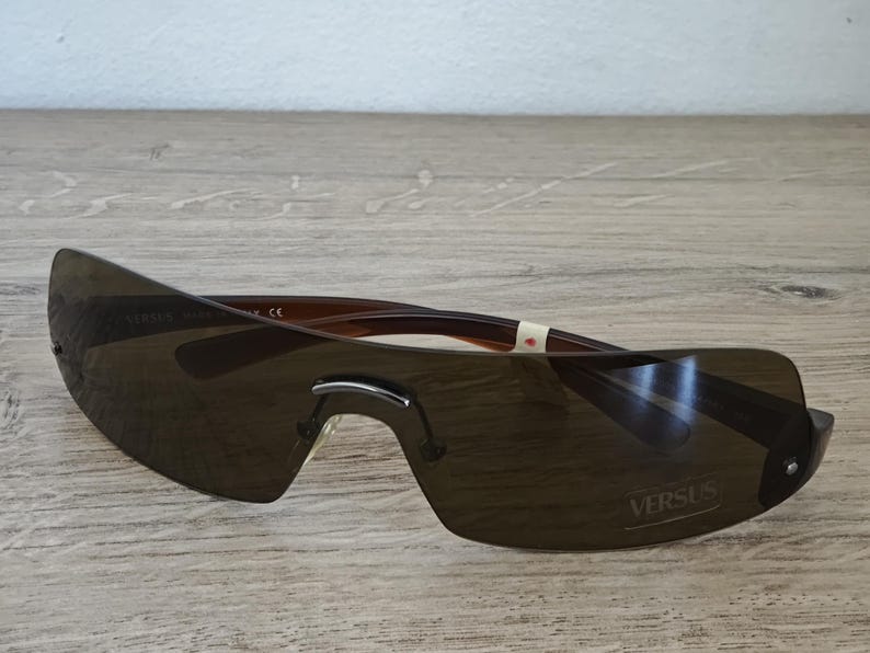 May include: Brown rimless sunglasses with dark tinted lenses. The sunglasses have a curved design and the word "VERSUS" is printed on the lens. The frame is brown and the lenses are a dark shade of brown.