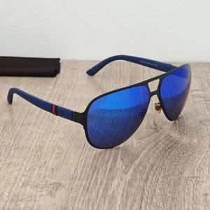 Puede incluir: Gafas de sol tipo aviador con montura negra mate y lentes de espejo azul vibrante. Las patillas presentan un patrón texturizado con un detalle de rayas rojas y verdes. El logotipo de Gucci está grabado en la esquina superior del cristal derecho.