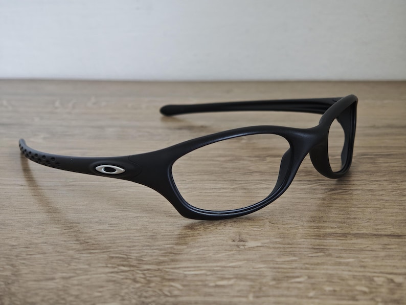 May include: Black eyeglasses with a sleek, modern design. The frames are matte black with a subtle curve and a small silver logo on the temple. The lenses are clear, and the arms have a perforated design for added style.