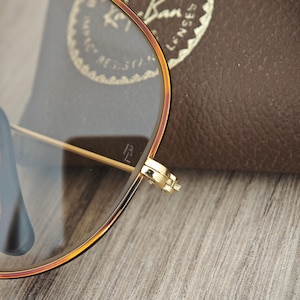 K&ouml;nnte beinhalten: Nahaufnahme einer goldgerahmten Brille mit einem braunen Lederetui im Hintergrund. Die Brille hat eine klassische Aviator-Form mit klaren Gl&auml;sern. Der Rahmen hat eine Farbverlauf, der von Gold zu einem r&ouml;tlich-braunen Farbton &uuml;bergeht.