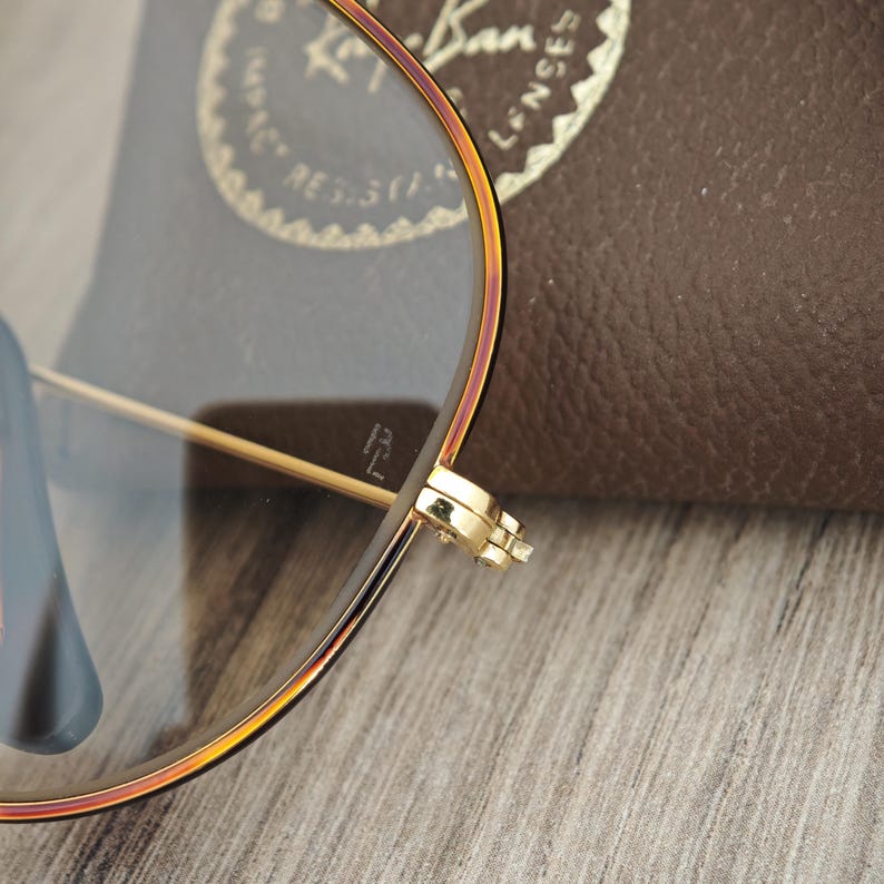 K&ouml;nnte beinhalten: Nahaufnahme einer goldgerahmten Brille mit einem braunen Lederetui im Hintergrund. Die Brille hat eine klassische Aviator-Form mit klaren Gl&auml;sern. Der Rahmen hat eine Farbverlauf, der von Gold zu einem r&ouml;tlich-braunen Farbton &uuml;bergeht.