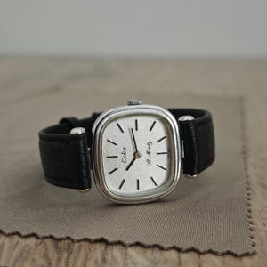 May include: A silver-toned Eska St. Moritz wristwatch with a square face and black leather band. The watch face is silver with black hands and hour markers. The band is black leather.