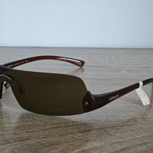 May include: Brown Versus sunglasses with a curved, shield-style lens. The frames are a dark brown color, with the word "VERSUS" printed on the temples. The sunglasses are made in Italy.