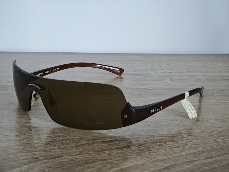 May include: Brown Versus sunglasses with a curved, shield-style lens. The frames are a dark brown color, with the word "VERSUS" printed on the temples. The sunglasses are made in Italy.