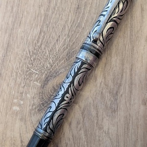 以下が含まれることがあります： 銀と黒の万年筆。装飾的なフローラルデザインが施されています。ペン先とキャップは黒、クリップはシルバーです。ペンの本体には詳細なフローラルパターンが刻まれています。木製の表面に表示されています。