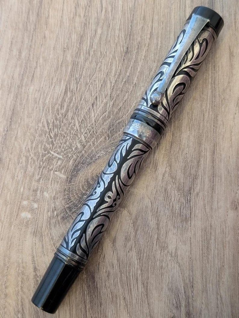 以下が含まれることがあります： 銀と黒の万年筆。装飾的なフローラルデザインが施されています。ペン先とキャップは黒、クリップはシルバーです。ペンの本体には詳細なフローラルパターンが刻まれています。木製の表面に表示されています。