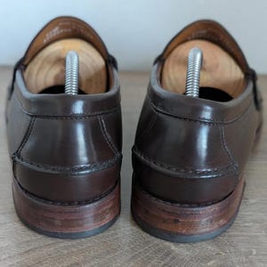 May include: A pair of brown leather loafers with wooden shoe trees inserted. The shoes have a glossy finish and a classic design. The soles are made of a darker brown material. The shoes are displayed on a light brown surface.