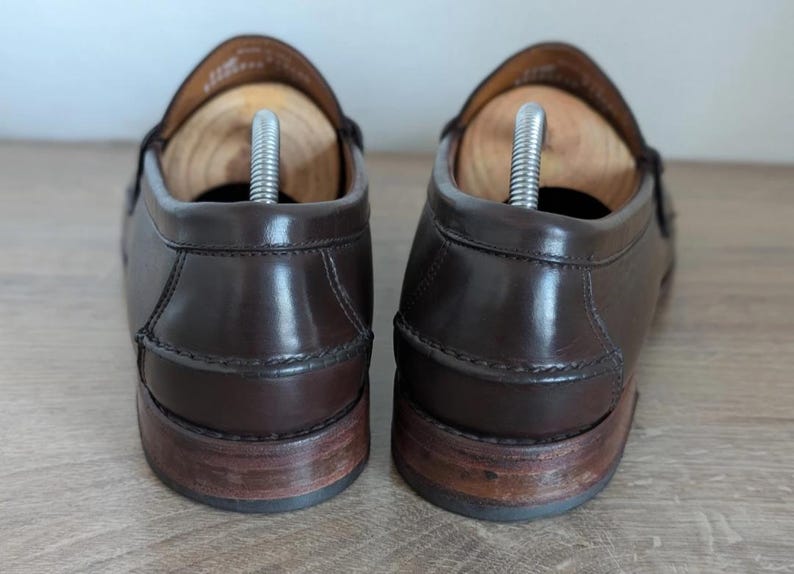 May include: A pair of brown leather loafers with wooden shoe trees inserted. The shoes have a glossy finish and a classic design. The soles are made of a darker brown material. The shoes are displayed on a light brown surface.