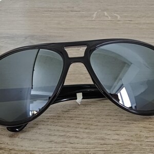 Puede incluir: Gafas de sol aviador negras con lentes reflectantes. Las gafas tienen una montura negra gruesa y un doble puente. Las lentes son de color plateado espejado, reflejando el entorno. El logotipo de Ray-Ban es visible en la lente izquierda.