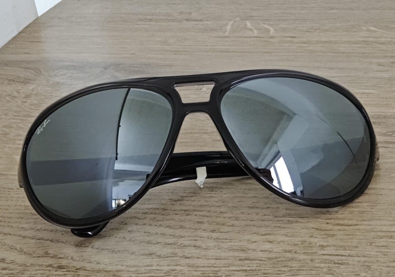 Puede incluir: Gafas de sol aviador negras con lentes reflectantes. Las gafas tienen una montura negra gruesa y un doble puente. Las lentes son de color plateado espejado, reflejando el entorno. El logotipo de Ray-Ban es visible en la lente izquierda.