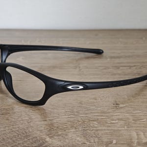 May include: Black sports eyeglasses with a sleek, modern design. The glasses feature a curved frame and temples, with the Oakley logo on the side. The lenses are clear, and the frame is made of a matte black material.