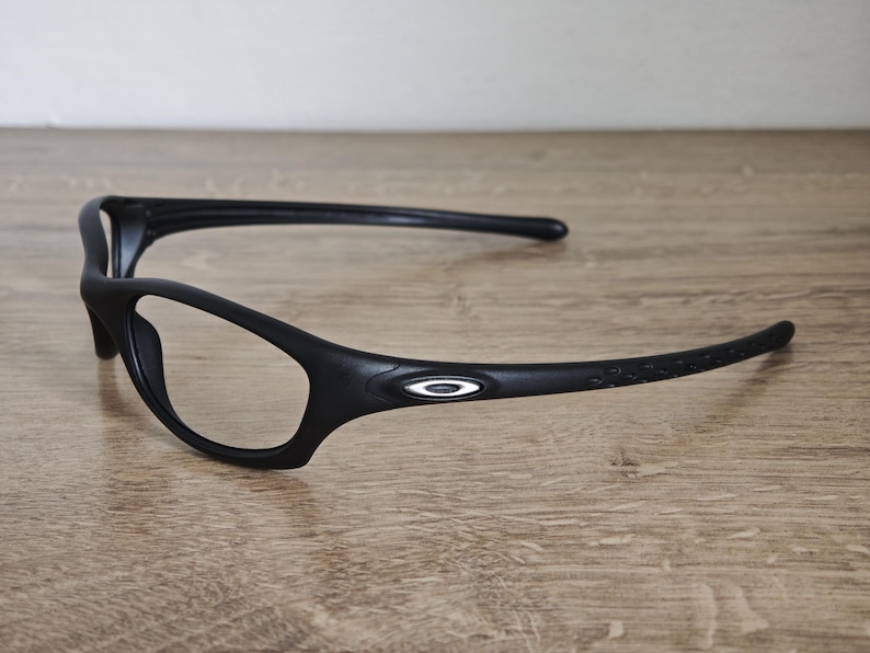 May include: Black sports eyeglasses with a sleek, modern design. The glasses feature a curved frame and temples, with the Oakley logo on the side. The lenses are clear, and the frame is made of a matte black material.