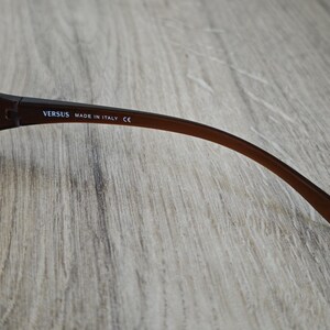 May include: Brown Versus sunglasses with a curved arm. The text "VERSUS MADE IN ITALY CE" is printed on the arm. The sunglasses have a silver lens and a brown frame.