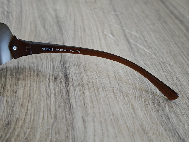May include: Brown Versus sunglasses with a curved arm. The text "VERSUS MADE IN ITALY CE" is printed on the arm. The sunglasses have a silver lens and a brown frame.