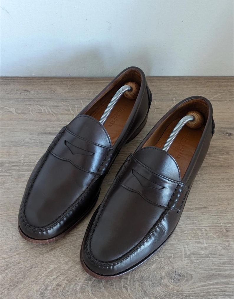 May include: A pair of dark brown leather penny loafers. The shoes have a classic design with a stitched vamp and a leather sole. Shoe trees are inserted in each shoe to maintain their shape. The loafers are displayed on a wooden surface.