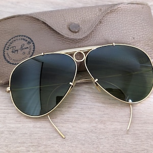 K&ouml;nnte beinhalten: Goldfarbene Aviator-Sonnenbrille mit dunkelgr&uuml;nen Gl&auml;sern, neben einem hellbraunen Lederetui. Das Etui hat einen Knopfverschluss und ein Logo "Bausch & Lomb Ray-Ban".