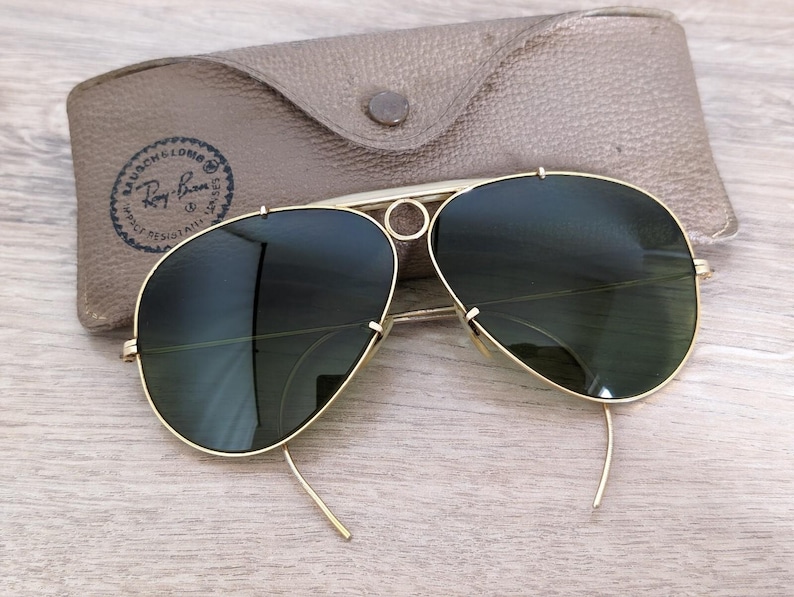 K&ouml;nnte beinhalten: Goldfarbene Aviator-Sonnenbrille mit dunkelgr&uuml;nen Gl&auml;sern, neben einem hellbraunen Lederetui. Das Etui hat einen Knopfverschluss und ein Logo "Bausch & Lomb Ray-Ban".
