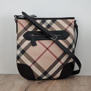 May include: A beige and brown plaid crossbody bag with a black leather strap and accents. The bag features a zippered pocket and a buckle closure. The bottom corners are black.