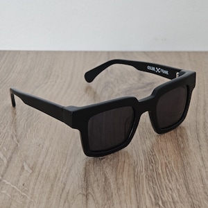 Könnte beinhalten: Schwarze Sonnenbrille mit einem dicken, quadratischen Rahmen. Die Sonnenbrille hat dunkle Gläser und der Text "OSCAR X FRANK" ist auf dem rechten Bügel aufgedruckt. Die Sonnenbrille liegt auf einer Holzoberfläche.