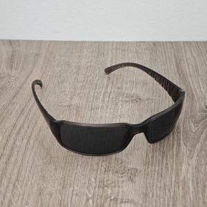 Puede incluir: Gafas de sol de color marr&oacute;n oscuro con un dise&ntilde;o de montura envolvente y lentes tintadas oscuras. Las patillas muestran letras en relieve a lo largo del lateral, y la estructura est&aacute; hecha de un material pl&aacute;stico liso y moldeado con forma curva.