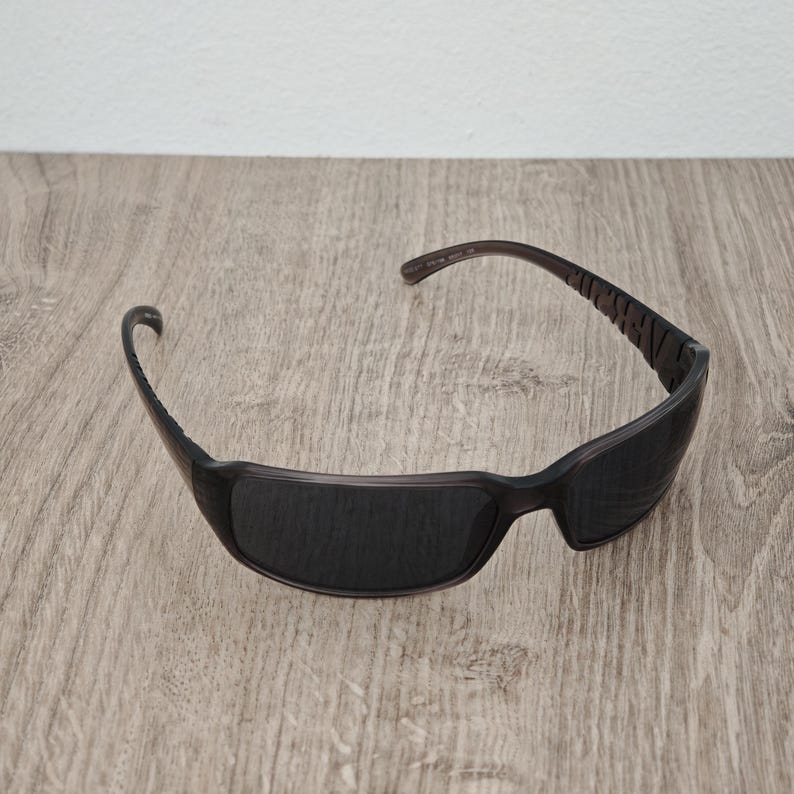 Puede incluir: Gafas de sol de color marr&oacute;n oscuro con un dise&ntilde;o de montura envolvente y lentes tintadas oscuras. Las patillas muestran letras en relieve a lo largo del lateral, y la estructura est&aacute; hecha de un material pl&aacute;stico liso y moldeado con forma curva.
