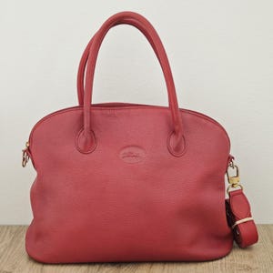 May include: A red leather handbag with a top handle and a detachable shoulder strap. The bag has a rounded shape, a zipper closure, and a small logo on the front. The hardware is gold-toned.