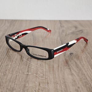 Peut inclure: Lunettes Dolce & Gabbana avec une monture rectangulaire noire et des branches aux motifs rouge, blanc et noir. Le nom de la marque Dolce & Gabbana est imprimé sur le verre droit et le logo D&G est visible sur la face interne de la branche.