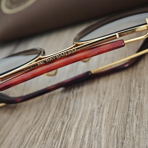 K&ouml;nnte beinhalten: Nahaufnahme einer Ray-Ban-Brille mit goldenen Rahmen und braunen B&uuml;geln. Der Text "B&L RAY-BAN USA" ist auf dem Rahmen zu sehen. Die Brille liegt auf einer Holzoberfl&auml;che.