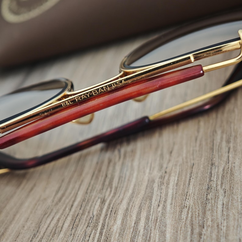K&ouml;nnte beinhalten: Nahaufnahme einer Ray-Ban-Brille mit goldenen Rahmen und braunen B&uuml;geln. Der Text "B&L RAY-BAN USA" ist auf dem Rahmen zu sehen. Die Brille liegt auf einer Holzoberfl&auml;che.