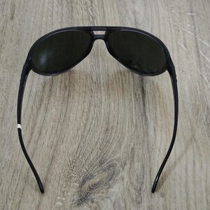 Puede incluir: Gafas de sol de aviador negras con lentes oscuras. Las gafas tienen un dise&ntilde;o elegante con un puente que conecta las dos lentes y patillas curvas. Las gafas est&aacute;n sobre un fondo de madera.