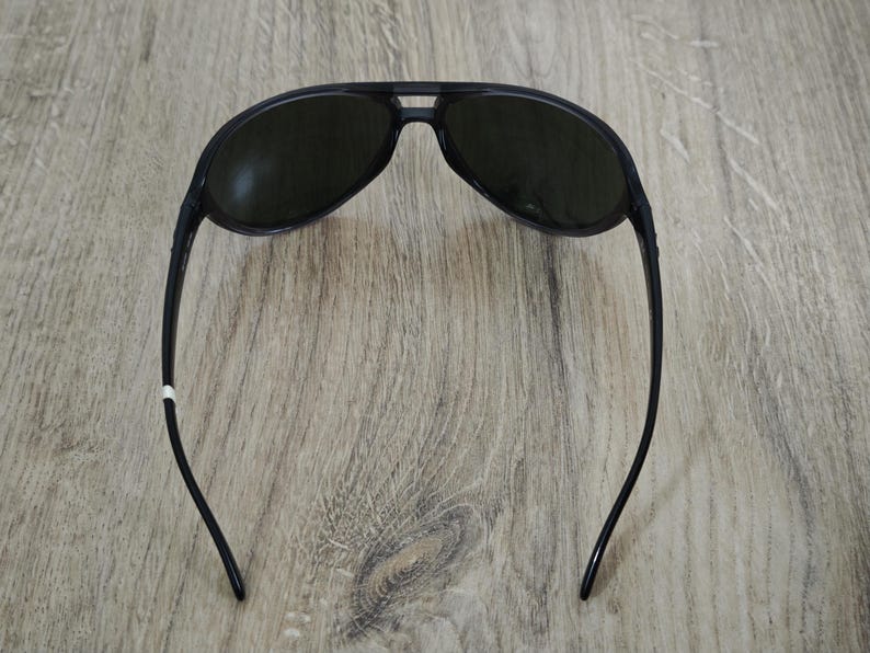 Puede incluir: Gafas de sol de aviador negras con lentes oscuras. Las gafas tienen un dise&ntilde;o elegante con un puente que conecta las dos lentes y patillas curvas. Las gafas est&aacute;n sobre un fondo de madera.