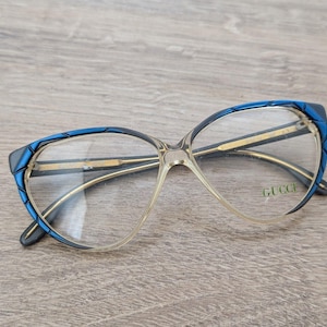 May include: Vintage eyeglasses with a clear frame and blue detailing. The cat-eye shaped glasses feature gold-toned arms and the brand name "GUCCI" on the lens. The glasses are set against a wooden background.