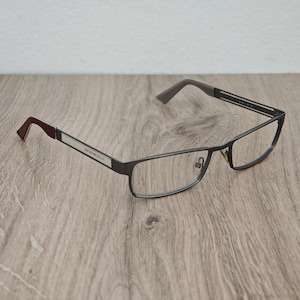 Può includere: Occhiali Emporio Armani con montatura in metallo grigio scuro e lenti rettangolari. Le aste presentano un inserto argentato con il nome del marchio, un terminale color bordeaux sul lato sinistro e un terminale grigio sul lato destro.
