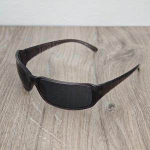 Puede incluir: Gafas de sol de color marr&oacute;n oscuro con montura rectangular y lentes oscuras. Las patillas presentan una superficie texturizada con patrones en los laterales, conectadas a un puente curvo y liso y a un frente que ofrece cobertura total.