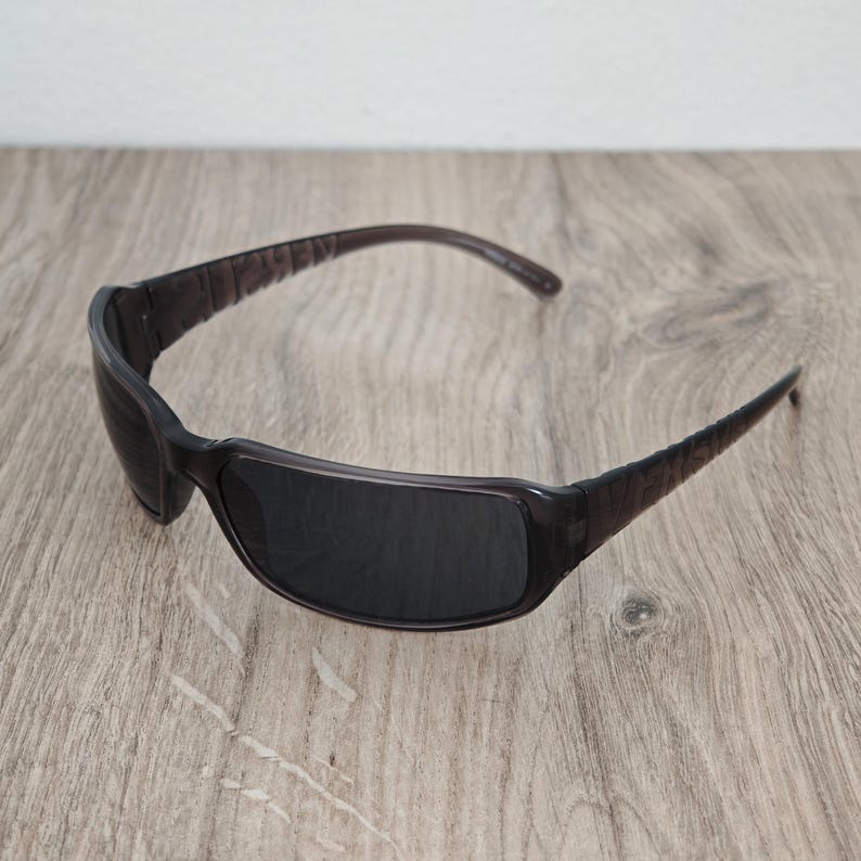Puede incluir: Gafas de sol de color marr&oacute;n oscuro con montura rectangular y lentes oscuras. Las patillas presentan una superficie texturizada con patrones en los laterales, conectadas a un puente curvo y liso y a un frente que ofrece cobertura total.