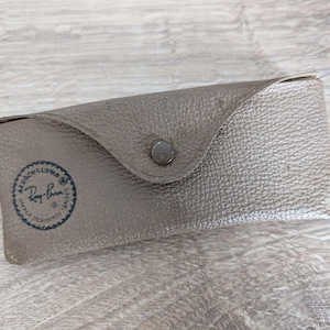 K&ouml;nnte beinhalten: Ein graues, strukturiertes Brillenetui mit einem Druckknopfverschluss. Das Etui hat eine abgerundete Klappe und ein rundes Logo mit der Aufschrift "Ray-Ban" in Dunkelblau. Das rechteckige Etui scheint aus Leder oder einem &auml;hnlichen Material zu bestehen.