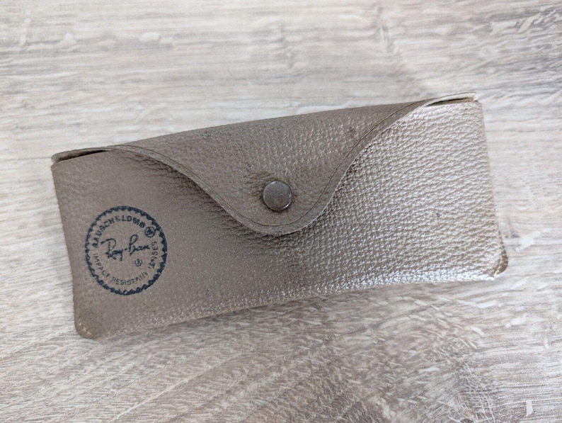 K&ouml;nnte beinhalten: Ein graues, strukturiertes Brillenetui mit einem Druckknopfverschluss. Das Etui hat eine abgerundete Klappe und ein rundes Logo mit der Aufschrift "Ray-Ban" in Dunkelblau. Das rechteckige Etui scheint aus Leder oder einem &auml;hnlichen Material zu bestehen.
