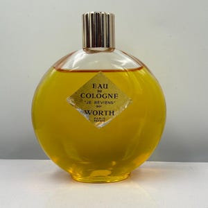 Könnte beinhalten: Eine Vintage-Flasche "Eau de Cologne" von Worth, mit einem goldfarbenen Verschluss und einem runden, gelb getönten Glaskörper. Das Etikett lautet "Je Reviens" und "Paris France". Die Flasche ist mit einer goldenen Flüssigkeit gefüllt.