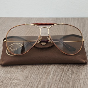 K&ouml;nnte beinhalten: Ray-Ban Aviator-Brille mit goldfarbenem Rahmen und braunen Schildpatt-Akzenten. Die Brille liegt auf einem braunen Lederetui mit dem Ray-Ban-Logo.