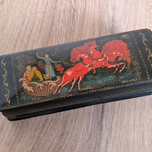 Può includere: Una scatola rettangolare nera laccata con un dipinto decorativo. Il dipinto raffigura una slitta trainata da cavalli con figure e un conducente. I cavalli sono rossi con grandi criniere stilizzate. La scatola ha un bordo dorato ornato.