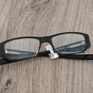 May include: A pair of Oakley Candidate 2.0 eyeglasses with a dark tortoise shell frame and clear lenses. The glasses are resting on a wooden surface. The text "Candidate 2.0" is visible on the frame.
