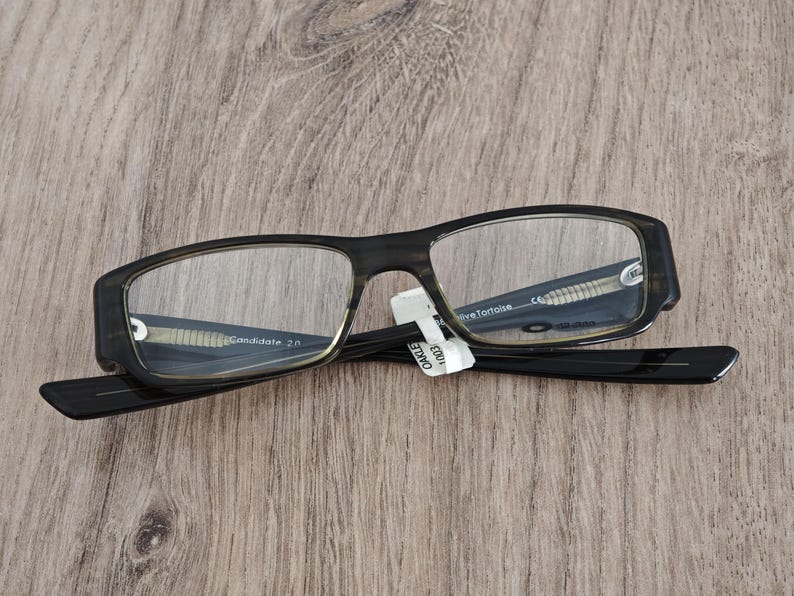 May include: A pair of Oakley Candidate 2.0 eyeglasses with a dark tortoise shell frame and clear lenses. The glasses are resting on a wooden surface. The text "Candidate 2.0" is visible on the frame.