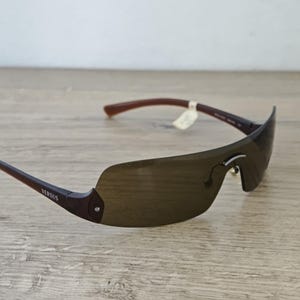 May include: Brown rimless sunglasses with dark lenses. The sunglasses have brown arms and the word "VERSUS" is printed on the left arm. The sunglasses are on a light brown wooden surface.