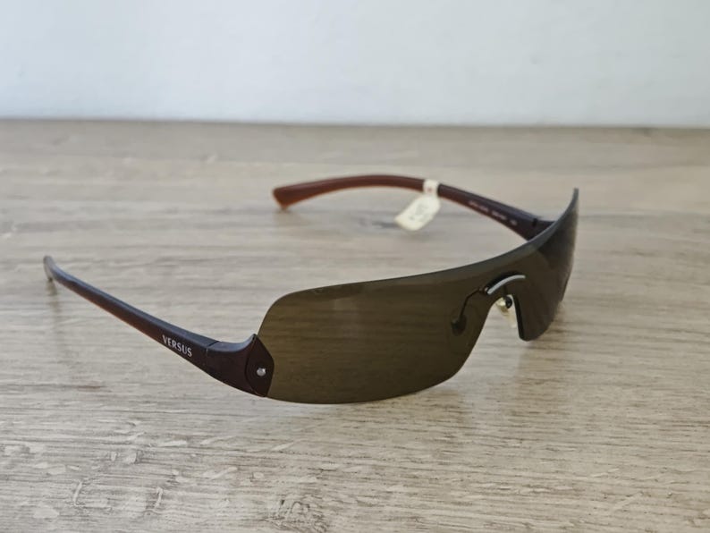 May include: Brown rimless sunglasses with dark lenses. The sunglasses have brown arms and the word "VERSUS" is printed on the left arm. The sunglasses are on a light brown wooden surface.